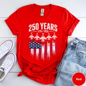 May include: A red t-shirt with a graphic design celebrating 250 years, from 1776 to 2026. The design includes the American flag and five white jet fighter silhouettes. The shirt is paired with white sneakers and blue jeans.