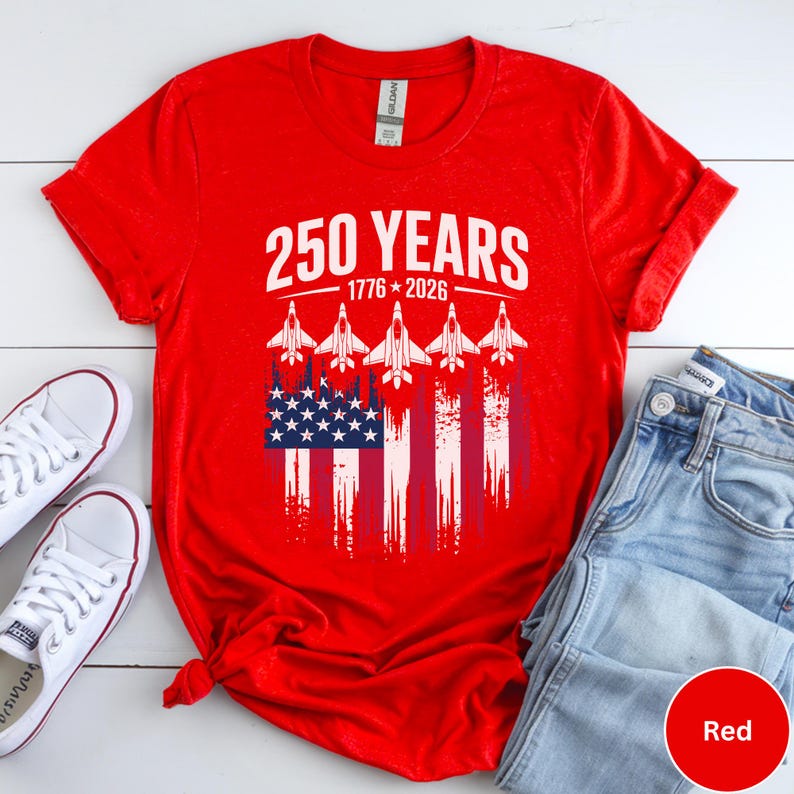 May include: A red t-shirt with a graphic design celebrating 250 years, from 1776 to 2026. The design includes the American flag and five white jet fighter silhouettes. The shirt is paired with white sneakers and blue jeans.