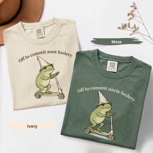 May include: Two Comfort Colors t-shirts, one ivory and one moss green, feature a whimsical frog design. The frog, wearing a wizard hat, rides a scooter. The ivory shirt reads "Off to commit aunt foolery," while the green shirt says "Off to commit uncle foolery."