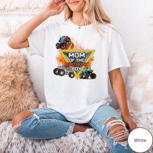 Puede incluir: Camiseta blanca con un gr&aacute;fico de tres monster trucks y el texto "MOM OF THE BIRTHDAY BOY" dentro de un tri&aacute;ngulo amarillo. Los camiones son rojos, plateados y verdes.