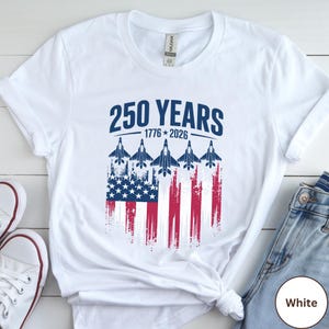 May include: White t-shirt featuring the text "250 YEARS 1776-2026" in navy blue. The design includes an American flag with jets flying above it. The shirt is a Gildan brand.