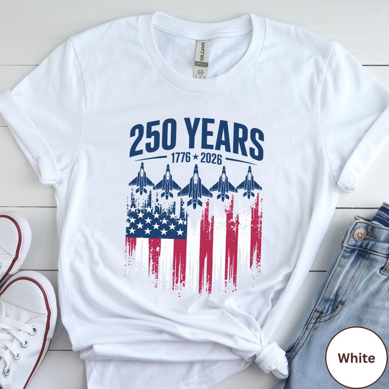 May include: White t-shirt featuring the text "250 YEARS 1776-2026" in navy blue. The design includes an American flag with jets flying above it. The shirt is a Gildan brand.