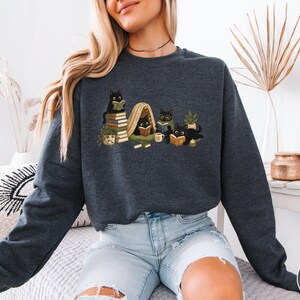 Puede incluir: Una sudadera de cuello redondo en gris oscuro jaspeado con un estampado gr&aacute;fico de cinco gatos negros leyendo libros. Los gatos est&aacute;n situados alrededor de pilas de libros, una peque&ntilde;a planta en maceta, una taza y una manta en tonos tierra.