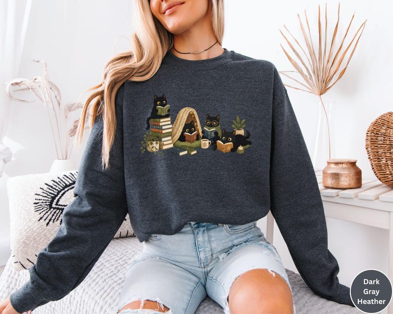 Puede incluir: Una sudadera de cuello redondo en gris oscuro jaspeado con un estampado gr&aacute;fico de cinco gatos negros leyendo libros. Los gatos est&aacute;n situados alrededor de pilas de libros, una peque&ntilde;a planta en maceta, una taza y una manta en tonos tierra.