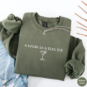 May include: Olive green sweatshirt with the text "a bride in a tini bit" and a martini glass graphic. A silver necklace with a small pendant is on the sweatshirt. The sweatshirt is folded over a pair of blue jeans.