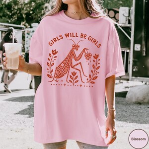 May include: A light pink t-shirt featuring a red graphic print of a praying mantis surrounded by floral elements and the text GIRLS WILL BE GIRLS arched above the insect. The design includes small star accents and a decorative border below.