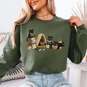 Puede incluir: Una sudadera de manga larga color verde militar con un estampado gr&aacute;fico de cuatro gatos negros leyendo libros. La ilustraci&oacute;n incluye gatos sentados sobre una pila de libros, dentro de una peque&ntilde;a tienda de campa&ntilde;a y tumbados, junto a plantas.