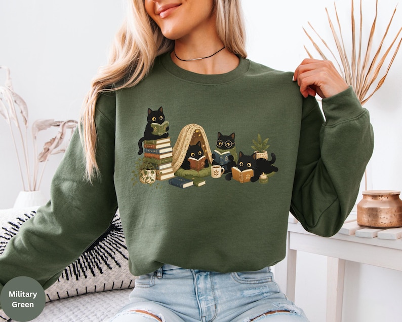 Puede incluir: Una sudadera de manga larga color verde militar con un estampado gr&aacute;fico de cuatro gatos negros leyendo libros. La ilustraci&oacute;n incluye gatos sentados sobre una pila de libros, dentro de una peque&ntilde;a tienda de campa&ntilde;a y tumbados, junto a plantas.