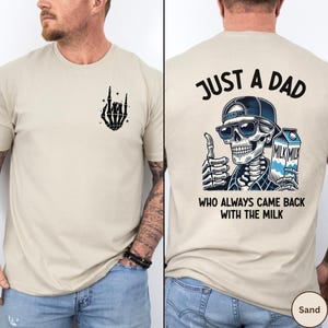May include: Sand-colored t-shirt featuring a black skeleton hand graphic on the front. The back of the shirt displays a skeleton wearing sunglasses and a cap, holding a milk carton, with the text "JUST A DAD WHO ALWAYS CAME BACK WITH THE MILK."