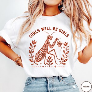 May include: A white t-shirt featuring a rust-coloured graphic of a praying mantis holding a small skull, surrounded by floral motifs and the text "GIRLS WILL BE GIRLS" arched above the insect. The design includes small star accents at the bottom.