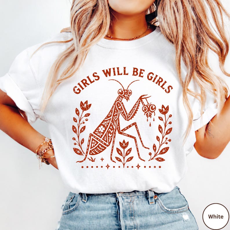 May include: A white t-shirt featuring a rust-coloured graphic of a praying mantis holding a small skull, surrounded by floral motifs and the text "GIRLS WILL BE GIRLS" arched above the insect. The design includes small star accents at the bottom.