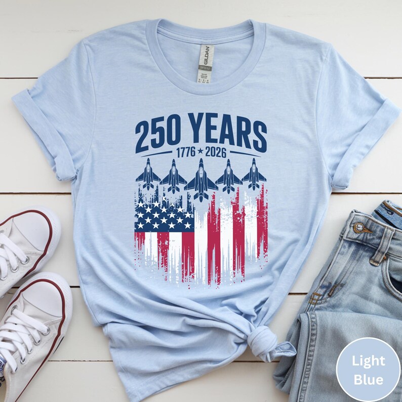 May include: Light blue t-shirt featuring the text "250 YEARS 1776-2026" above a graphic of the American flag with jets. The shirt is made of a soft material and is suitable for casual wear.