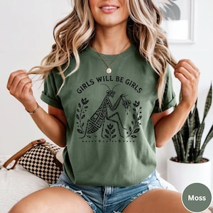 May include: A moss green short-sleeve t-shirt featuring a black graphic print of a praying mantis surrounded by floral stems. The text "GIRLS WILL BE GIRLS" is printed in a curved black font above the insect illustration on the front of the shirt.