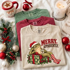 May include: Three crew neck t-shirts in beige, maroon, and green. The beige shirt features a graphic of a frog wearing a Santa hat, reclining on Christmas presents, holding a mug, with the text MERRY, APPARENTLY. The shirts have Comfort Colors labels.