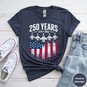 May include: Heather midnight blue t-shirt with the text "250 YEARS 1776-2026" above a graphic of the American flag and five white jets. The shirt is paired with white sneakers and blue jeans.