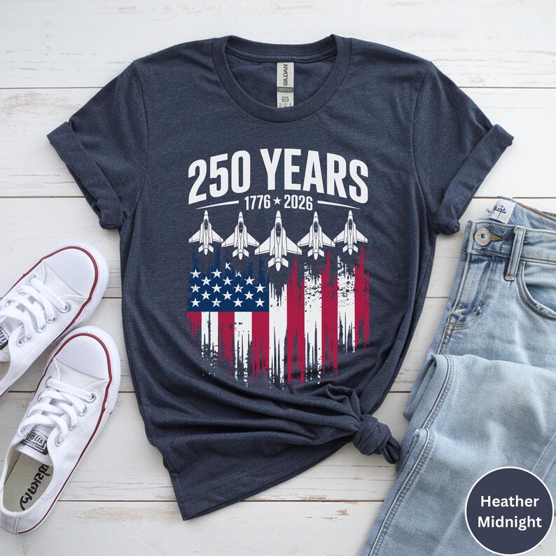 May include: Heather midnight blue t-shirt with the text "250 YEARS 1776-2026" above a graphic of the American flag and five white jets. The shirt is paired with white sneakers and blue jeans.