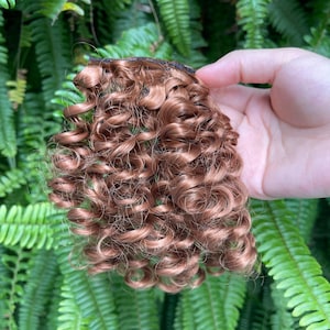 May include: A hair extension piece featuring tight, spiral curls in a warm auburn brown shade. The hair is attached to a dark, horizontal weft strip at the top, designed for secure application. The texture is defined with distinct, bouncy ringlets.