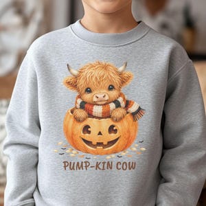 Könnte beinhalten: Ein hellgraues Sweatshirt mit Rundhalsausschnitt und einer Grafik eines flauschigen Hochlandrindes mit gestreiftem Schal, das in einem geschnitzten Kürbis sitzt. Der Text PUMP-KIN COW ist in braunen Buchstaben unter dem Kürbis abgebildet.