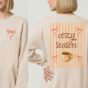 Könnte beinhalten: Ein sandfarbenes Sweatshirt mit Rundhalsausschnitt und einer kleinen orangefarbenen Schleifengrafik auf der linken Brust. Die Rückseite zeigt vertikale orangefarbene Streifen, den Schriftzug "cozy season" und eine gelbe Tasse.