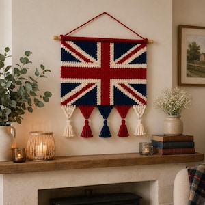 Puede incluir: Un tapiz rectangular que presenta un diseño de bandera Union Jack de ganchillo en rojo, blanco y azul oscuro. La pieza está suspendida de un pasador de madera con un cordón rojo y terminada con cinco borlas de hilo alternas en crema y rojo.