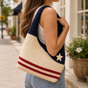 May include: A crochet shoulder bag featuring a color-blocked design with a dark navy blue upper section, a cream-colored lower section, and two horizontal red stripes near the bottom. A small cream-colored star is stitched onto the navy blue area.