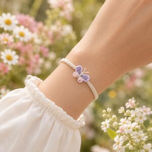 May include: A delicate macrame bracelet featuring a small, central butterfly charm crafted from light purple and soft pink thread. The bracelet band is made of a neutral beige cord, showcasing a woven texture that encircles the wrist.