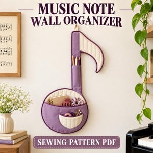 Peut inclure: Un organisateur mural en tissu en forme de note de musique dans des tons violet et crème. Il comporte une poche verticale haute pour des crayons et une grande base circulaire avec deux poches supplémentaires contenant des petits objets et un carnet.