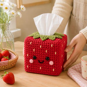 Puede incluir: Una funda de ganchillo para caja de pañuelos con forma de fresa roja y hojas verdes en la parte superior. La fresa tiene una cara sonriente con ojos de cuentas negras, boca bordada y mejillas rosadas, con pañuelos blancos en el centro.