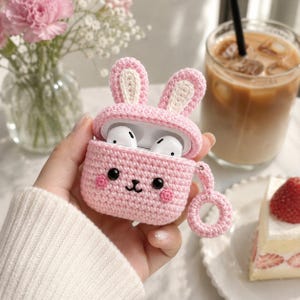 May include: A light pink crocheted protective case for wireless earbuds, featuring two upright rabbit ears with cream-colored centers, black bead eyes, a stitched nose and mouth, and two small pink circular cheeks, with a matching crocheted ring loop.