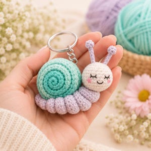 Puede incluir: Un llavero de caracol tejido a ganchillo con una concha en espiral de color verde menta, un cuerpo morado claro y una cabeza color crema con ojos cerrados bordados, una pequeña sonrisa y mejillas rosadas. Unido a una anilla de metal plateado.