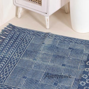 May include: A rectangular blue and white rug featuring a central geometric pattern of alternating striped squares, bordered by a braided design and fringe on the short edge. The text "AjenWeave" is printed in a dark font on the surface of the rug.