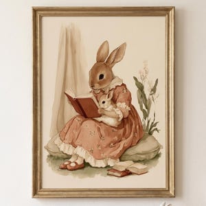 May include: A framed wall art print featuring a brown rabbit wearing a floral dress, sitting on a cushion while reading a book and holding a smaller white rabbit. The illustration uses soft earth tones and includes two additional books on the ground.