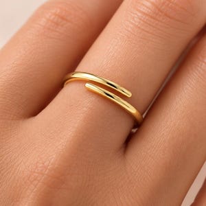 以下が含まれることがあります： 2つの丸みを帯びた端が重なり合うオープンバイパスデザインの光沢のあるゴールドトーンのリング。バンドは滑らかで反射する表面を持ち、指を部分的に包み込む細く連続したプロファイルが特徴です。