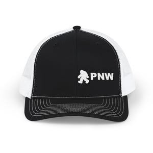 May include: A black and white trucker cap featuring a solid black front panel with a white embroidered graphic of a Bigfoot silhouette next to the text PNW. The cap includes a white mesh back, a black brim with stitching, and a black button on top.