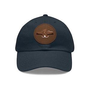May include: A navy blue baseball cap featuring a circular brown leather patch on the front. The patch displays an embossed graphic of a walking Bigfoot silhouette set against a backdrop of a mountain range and a forest of pine trees.