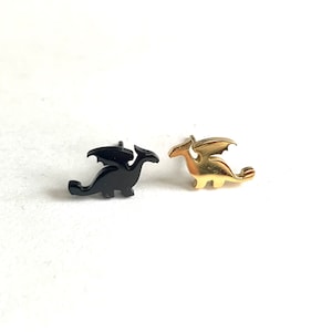 May include: Two dragon-shaped stud earrings. One is black, the other is gold-coloured. The dragons have outstretched wings and are facing to the right. The earrings are on a white background.