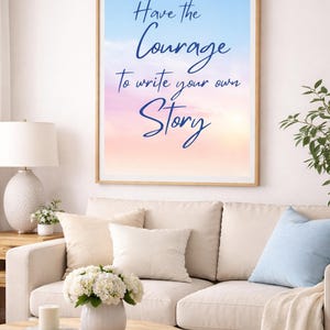 May include: A framed print with the text "Have the Courage to write your own Story" in blue script against a pastel sky background. The print hangs above a cream-coloured sofa with decorative cushions and a wooden coffee table.