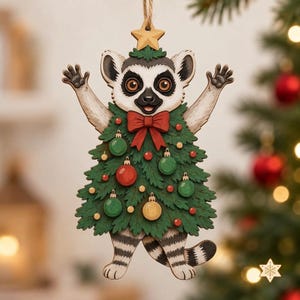 Könnte beinhalten: Ein hölzerner Lemuren-Anhänger mit erhobenen Armen, der ein grünes Weihnachtsbaumkostüm trägt, verziert mit roten, grünen und goldenen Kugeln sowie einer großen roten Schleife. Ein kleiner Holzstern krönt die Figur, befestigt an einer braunen Schnur.