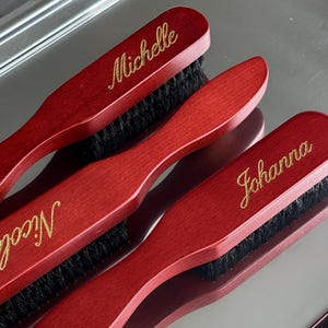Könnte beinhalten: Drei personalisierte rote Holzbürsten mit schwarzen Borsten. Die Namen "Michelle", "Nicole" und "Johanna" sind in goldener Schrift auf jede Bürste geschrieben. Die Bürsten sind auf einer reflektierenden silbernen Oberfläche angeordnet.
