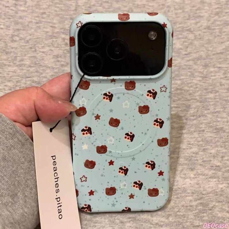 May include: A light blue phone case with a repeating pattern of brown bear faces, stars, and cake slices. The case has a camera cutout and a white circular area on the back. The brand name "peaches.pitao" is on a tag.