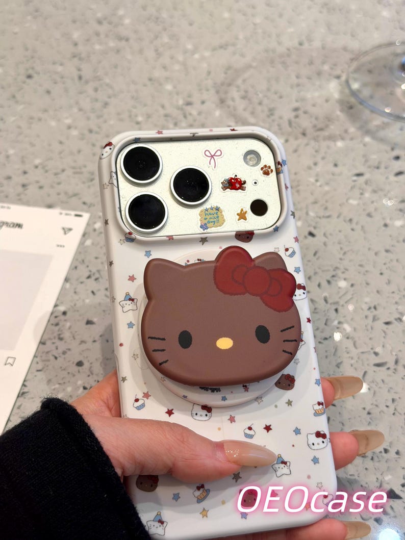 Puede incluir: Funda de tel&eacute;fono blanca con un dise&ntilde;o de Hello Kitty marr&oacute;n y un lazo rojo, adem&aacute;s de un agarre desplegable. La funda tiene un estampado de estrellas, cupcakes y caras de Hello Kitty. El texto "OEOcase" es visible.