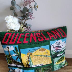 May include: Green cushion featuring the word "QUEENSLAND" in red. The design includes images of a lighthouse, a boat, and a landscape. The words "TROPICAL NORTH" are also visible.