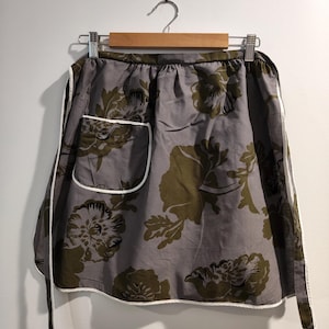 May include: A waist apron featuring a grey base colour with a large-scale floral pattern in olive green and dark grey. The apron includes a single patch pocket on the right side, finished with white piping along the edges and a white waist tie.