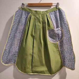 May include: A pleated apron featuring a solid olive green centre panel and light blue side panels with a white swirling vine pattern. The apron includes a single patch pocket on the right side, finished with light yellow piping along the edges.