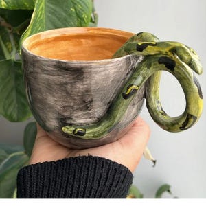 May include: A ceramic mug with a gray, brushed-texture exterior and a smooth, orange interior. The handle is shaped like a green snake with black and yellow markings that wraps around the side of the mug, with the head resting near the rim.