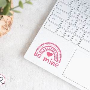 May include: A pink and white rainbow sticker with a heart in the center and the text "Be mine" on a white laptop.