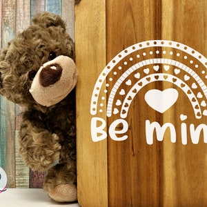 May include: A brown teddy bear peeking around a wooden sign with a white rainbow design and the text "Be mine".