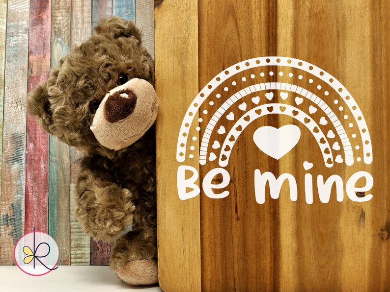 May include: A brown teddy bear peeking around a wooden sign with a white rainbow design and the text "Be mine".