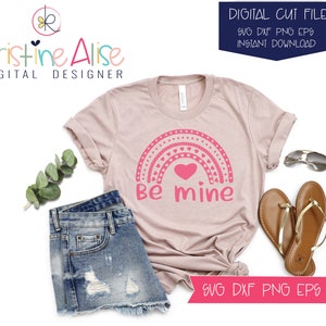 May include: A pink and white rainbow with hearts and the text "Be mine" on a light pink t-shirt. The t-shirt is on a white background with a pair of sunglasses, a pair of sandals, and a pair of denim shorts.