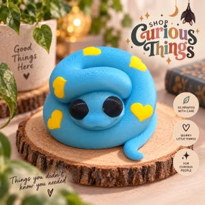 May include: A bright blue 3D printed coiled snake figurine featuring smooth yellow heart-shaped patterns across its body and two large, round, solid black eyes. The snake is positioned in a curled, resting posture on a circular wooden slice.